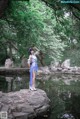 A woman in a blue dress standing on a rock by a pond.